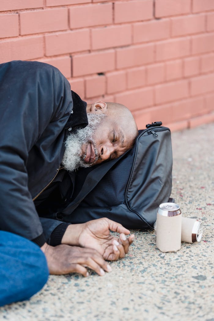 cta-02 An elderly man rests on a sidewalk, leaning against a brick wall with a can beside him.